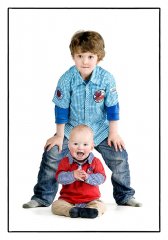 Kinderfotografie in studio van baby in het rood zit en zijn trotse broertje in het blauw staat erachter met zijn benen gespreid gefotografeerd door fotograaf in de fotostudio. www.marijnissenfotografie.nl
